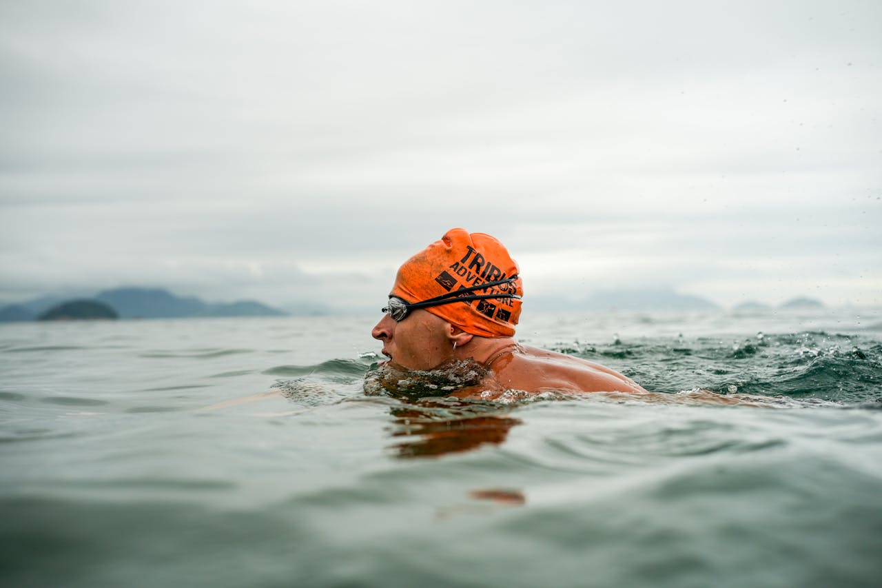 Adult male swimmer in open water wearing an orange swim cap and goggles, swimming in the sea.